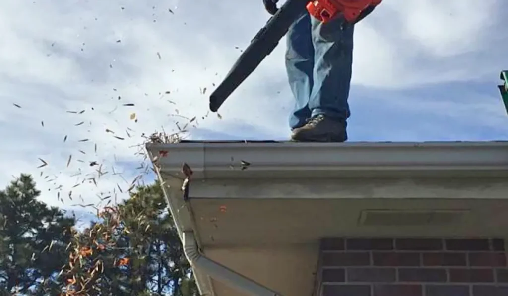 A professional gutter cleaning company worker standing on a roof, using a leaf blower to remove gutter debris.