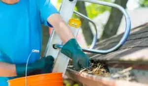 A professional gutter cleaner removing debris from a roof gutter using gloves and a bucket.