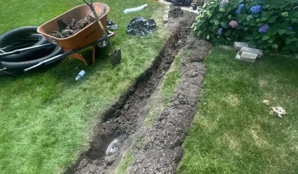 Open trench in backyard showing underground drainage system installation with visible pipes, tools, and soil removed.