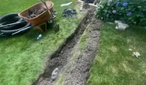 Open trench in backyard showing underground drainage system installation with visible pipes, tools, and soil removed.